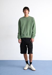 Grüner Sweatshirt mit Rundhalsausschnitt und gerippten Bündchen, kombiniert mit schwarzen Cargo-Shorts und weißen Sportschuhen. Einfaches Design, lässiger Schnitt.
