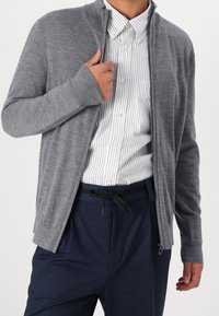 Šedý zip-up svetr s ribovaným límcem a manžetami, nošený přes bílou pruhovanou košili s knoflíky, doplněný tmavě modrými kalhotami.