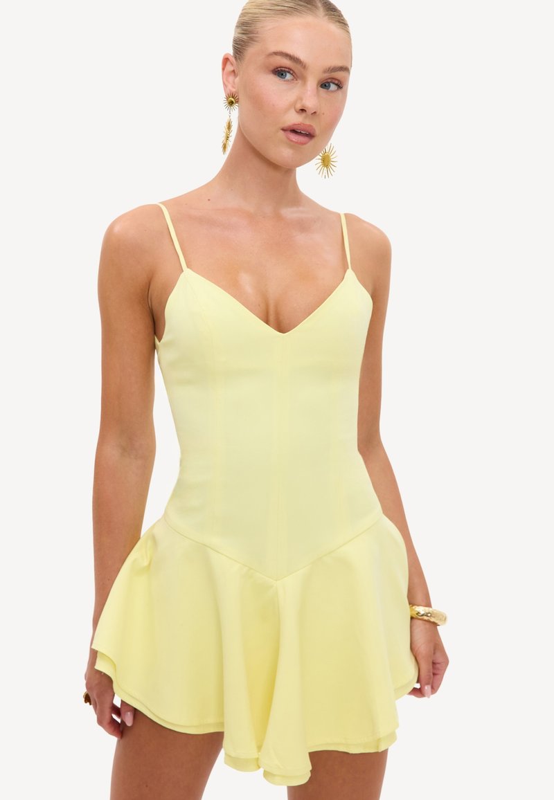 Femme portant une robe sans manches jaune clair avec des fines bretelles et une jupe évasée, accessoirisée de boucles d’oreilles dorées en forme de soleil et d’un bracelet en or.
