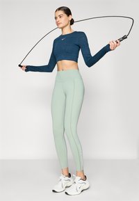 Világoszöld leggings varrási részletekkel és logóval, sötétkék cropped felsővel, amely texturált mintázattal rendelkezik, és fehér sportcipővel.