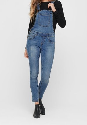 Mujer con peto vaquero azul ajustado combinado con una camiseta negra de manga larga y botines negros con tachuelas plateadas.