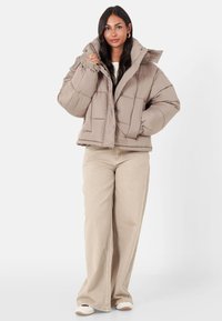 Beige pufferjacka med en kort design, med hög krage, knappstängning och kviltad yta. Matchas med ljust beigefärgade vida byxor.