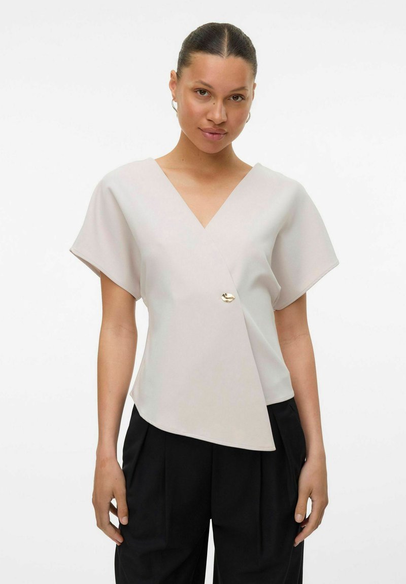 Lys beige omslagsbluse med V-hals, korte ermer og asymmetrisk skjæring, med et gullfarget knappeakcent foran.