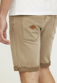 Beige Shorts mit umgeschlagenen Säumen, ausgestattet mit einem Leder-Diamant-Patch auf der Gesäßtasche. Hergestellt aus Denim mit einer glatten, weichen Textur.