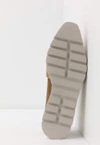 Semelle d'une chaussure en cuir suédé beige avec une semelle extérieure en caoutchouc épais et strié, adossée à un mur et un sol blancs.