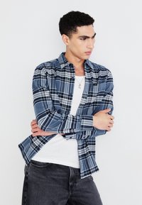 Camisa de franela de cuadros azules con patrones en blanco y azul marino, con diseño de botones, usada sobre una camiseta sin mangas blanca y combinada con jeans oscuros.
