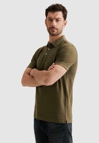 Hombre con cabello corto y castaño y barba, vistiendo una camiseta polo verde oliva y jeans oscuros, de pie con los brazos cruzados contra un fondo gris.