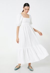 Vestido de algodón blanco con escote cuadrado, mangas abullonadas y dobladillo en capas. Combinado con zapatos planos en tono azul verdoso para un contraste. Tela simple y texturizada.