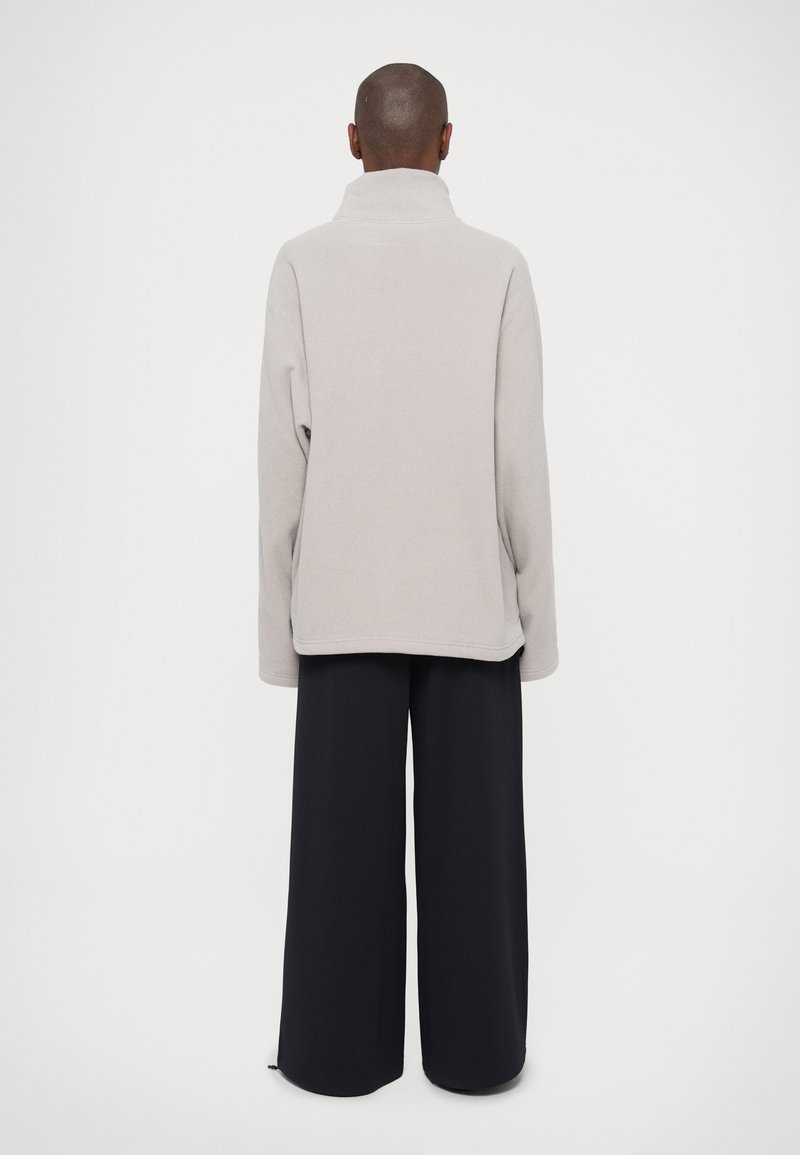 Persoon in een lichtgrijze coltrui en wijde zwarte broek, staande met het gezicht naar een effen witte achtergrond.