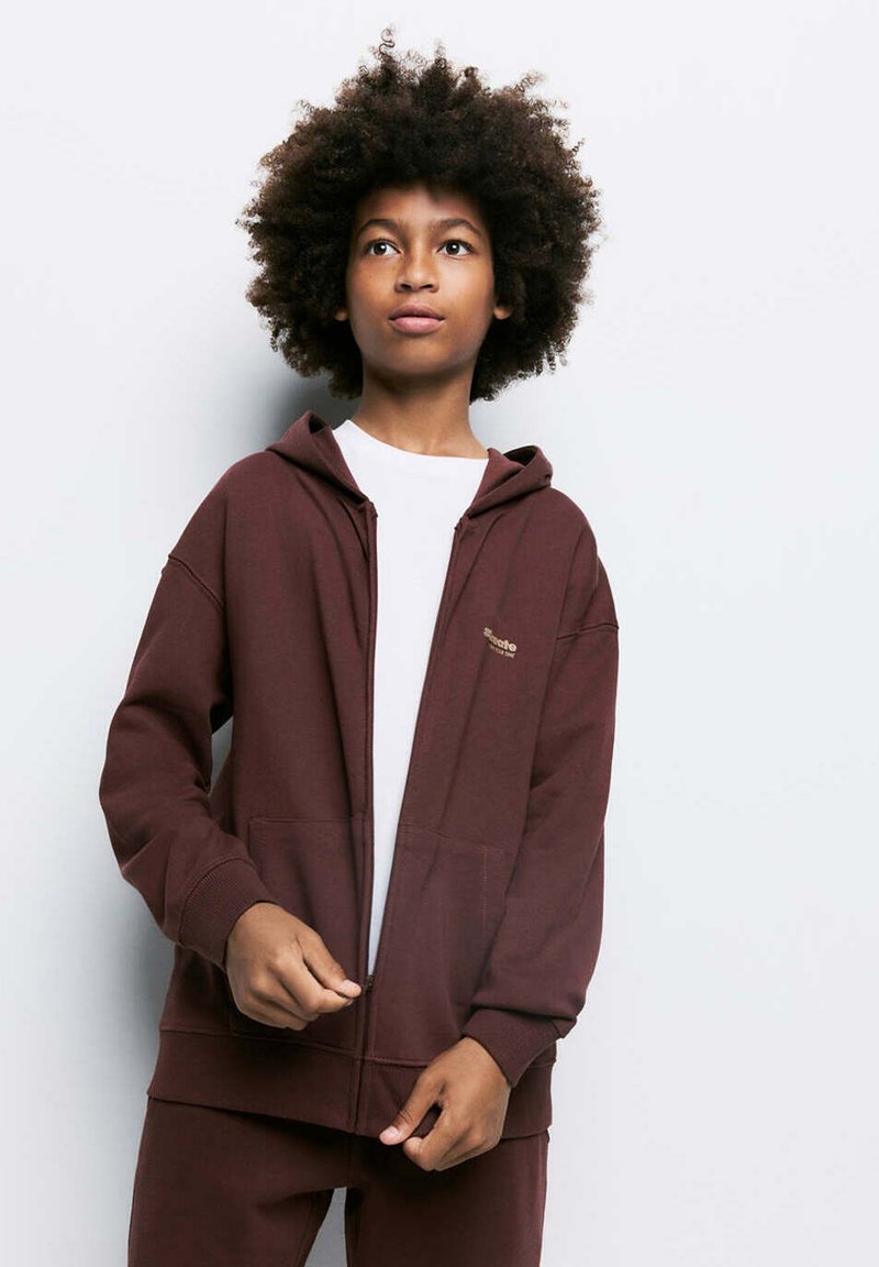 Garçon aux cheveux bouclés portant un sweat à capuche marron zippé et un pantalon assorti par-dessus une chemise blanche, debout devant un fond clair uni.