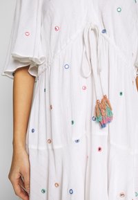 Robe blanche avec manches volantées, cercles brodés multicolores et un nœud devant orné de pompons roses, bleus, verts et oranges.