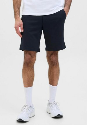 Homme portant un short noir jusqu'aux genoux, des chaussettes blanches, des baskets blanches et une chemise blanche, debout avec une main dans la poche.