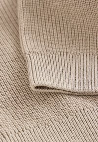 Tissu tricot beige avec une texture de côtes verticales, présentant un bord de poignet replié. Construction en fil fin, aspect doux et flexible.
