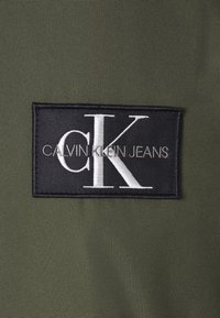 Štítek Calvin Klein Jeans s bílou vyšívanou „CK“ a „CALVIN KLEIN JEANS“ na černém obdélníkovém písmu na olivově zelené tkanině.