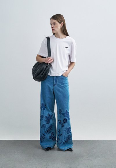 Άσπρο βαμβακερό t-shirt με μια μικρή μαύρη ετικέτα, σε συνδυασμό με ανοιχτό μπλε παντελόνι με φαρδιά πόδια που διαθέτει σκούρη μπλε κεντητική με λουλούδια και μια μαύρη τσάντα.