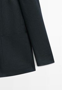 Tmavě šedý rukáv saka a detail přední kapsy na jednoduchém bílém pozadí.