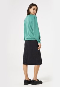 Sweatshirt vert menthe avec épaules bouffantes, associé à une jupe en denim noir. Le modèle porte des mocassins noirs à semelles épaisses, se tournant légèrement vers l'arrière.