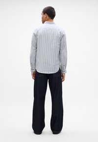 Gai pelēku svītrainu krekls ar garām piedurknēm, savienots ar tumši ziliem platām biksēm, skatīts no aizmugures.