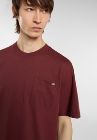 Burgundská bavlněná tričko s krátkým rukávem, kulatým výstřihem a přední kapsou s malou nášivkou. Hladká textura.