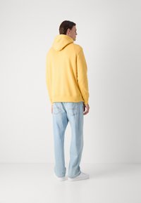 Sweatshirt à capuche jaune avec poche kangourou, associé à un jean bleu clair ample. Tissu lisse, design décontracté et baskets blanches simples.