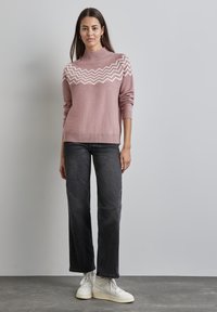 Rosa gestrickter Pullover mit hohem Kragen und weißem Zickzackmuster, kombiniert mit schwarzen weit geschnittenen Jeans und weißen Sneakers. Glatte Textur.