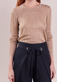 Suéter de manga larga con canalé en beige metálico, que presenta un escote redondo. Combinado con pantalones de tiro alto de color navy con un detalle de lazo y acentos metálicos.