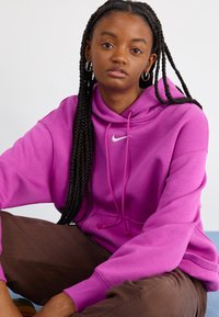 Μωβ oversized φούτερ με κορδόνι, με λευκό λογότυπο Nike. Το μοντέλο κάθεται με καφέ παντελόνι, δείχνοντας πλεξούδα στα μαλλιά.