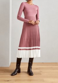 Pitkähihainen, istuva neulemekko vaaleanpunaisena, jossa on laskostettu valkoinen hameosa. Yksityiskohtina viininpunaiset raidat helmassa. Yhdistetty mustiin polveen saakka ulottuviin saappaisiin.