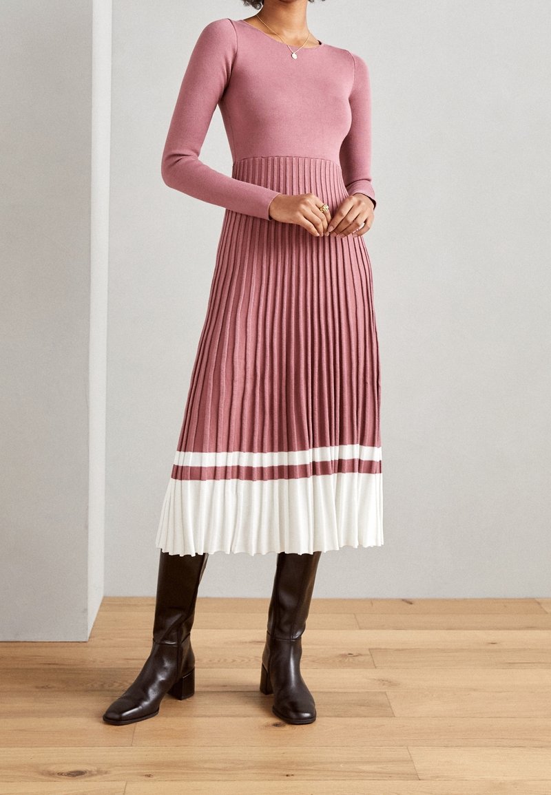 Pitkähihainen, istuva neulemekko vaaleanpunaisena, jossa on laskostettu valkoinen hameosa. Yksityiskohtina viininpunaiset raidat helmassa. Yhdistetty mustiin polveen saakka ulottuviin saappaisiin.