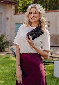 Black quilted clutch purse held in hand, featuring a geometric pattern. Model wears a short-sleeve white top and a burgundy skirt.