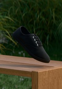 Schwarze Canvas-Sneaker mit Schnürsenkeln, die über einer Holzbank im Freien schweben, mit grünem Gras und Pflanzen im Hintergrund.