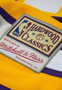 Hardwood Classics-Label auf einem gelben Sporttrikot, mit einem weißen und lila Rand, auf dem "Mitchell & Ness" in roter Schrift steht.