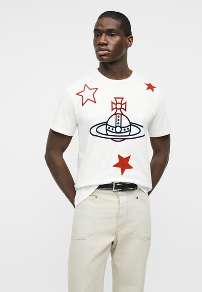 Άνδρας που φοράει λευκό μπλουζάκι με κόκκινα αστέρια και μια σκούρα μπλε σφαίρα σαν πλανήτης με σταυρωτό σχέδιο, συνδυασμένο με ανοιχτό μπεζ παντελόνι και ζώνη.