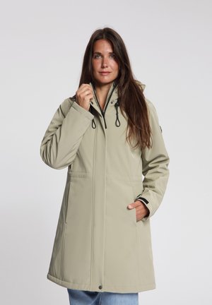 Vrouw met lang bruin haar die een beige jas met capuchon draagt, één hand in de zak en de andere die de rits bij de kraag verstelt, tegen een effen achtergrond.