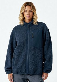 Veste en polaire bleu marine avec fermeture éclair à l'avant, col montant, poches latérales, et tissu texturé avec un accent en tissu lisse sur la poitrine.