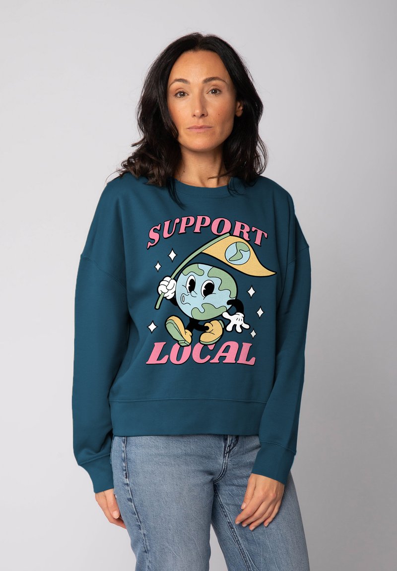 Oversized sweatshirt in blauwgroen met een cartoonachtig Aardebol-karakter met een vlag, de tekst "SUPPORT LOCAL" en lichte diamantaccenten in roze en geel.