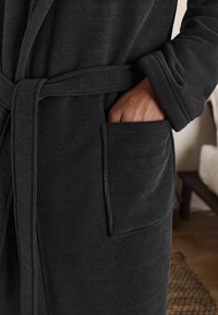 Rochie neagră din țesătură ribbed, cu talie legată, mâneci lungi și un buzunar frontal, având detalii texturate pe material.