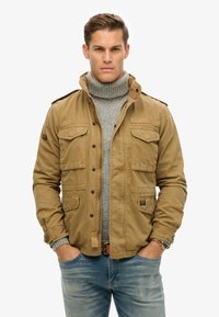 Khaki jas met meerdere voorzakken en een hoge kraag, gedragen over een grijze turtleneck. De jas heeft een gladde textuur en een knoopsluiting.