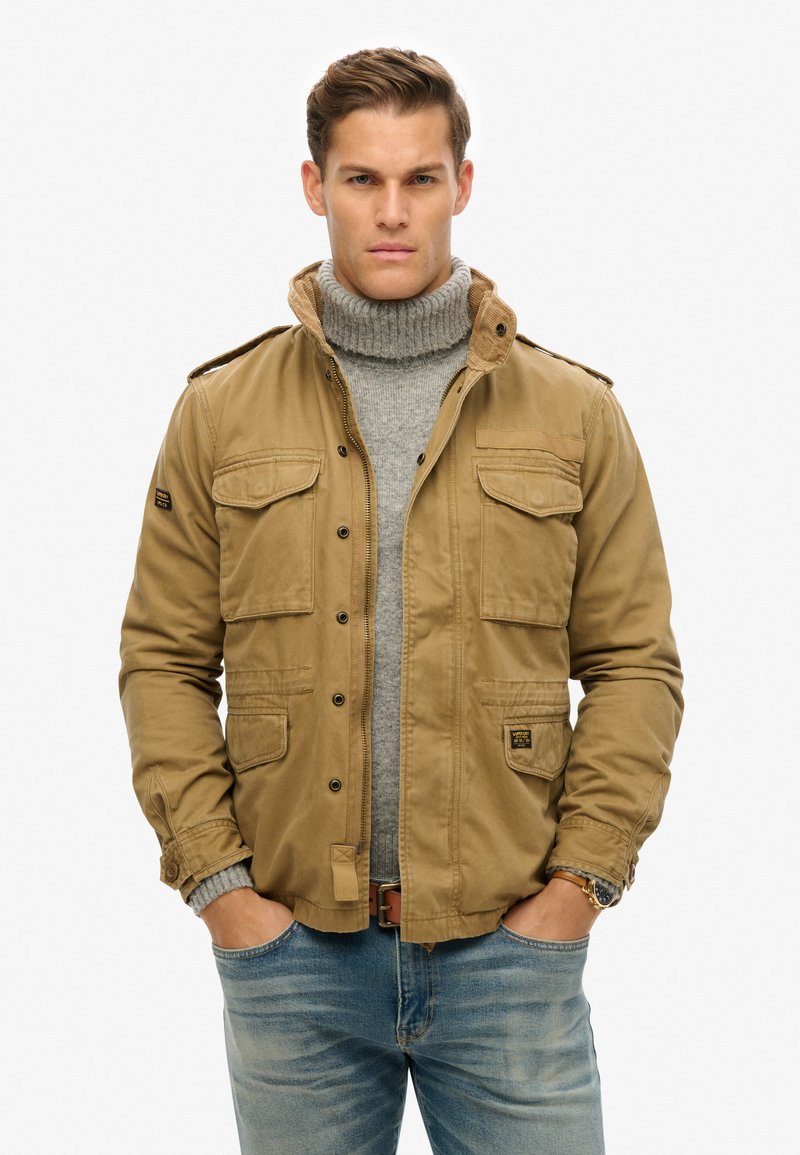 Khaki jas met meerdere voorzakken en een hoge kraag, gedragen over een grijze turtleneck. De jas heeft een gladde textuur en een knoopsluiting.