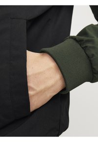 Giacca nera con polsino a coste verde, dotata di tasca sulla cucitura laterale. Il materiale sembra liscio con una leggera lucentezza. La mano è posizionata nella tasca.