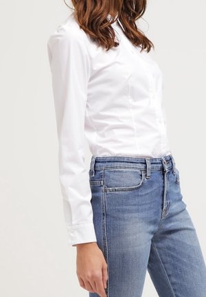 Donna con una camicia bianca aderente a maniche lunghe con bottoni, infilata in jeans di denim blu a vita alta, in piedi davanti a uno sfondo semplice.