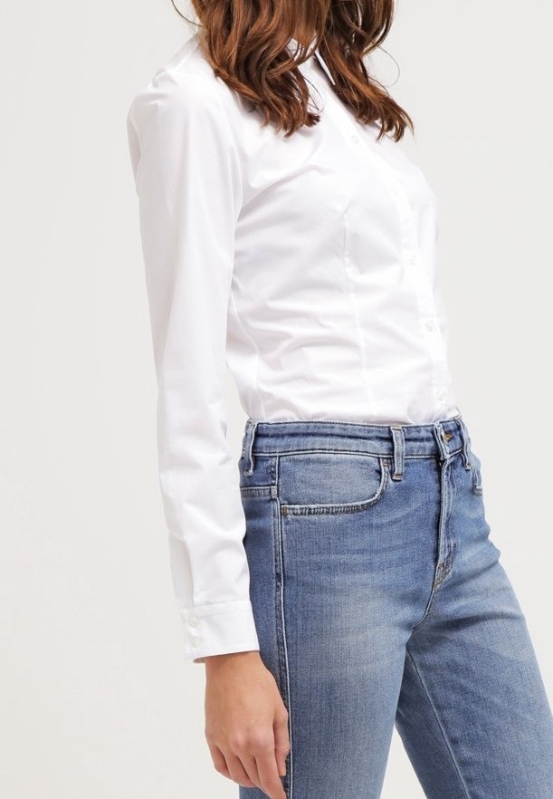 Femme portant une chemise blanche cintrée à manches longues boutonnée, rentrée dans un jean en denim bleu taille haute, debout devant un fond uni.