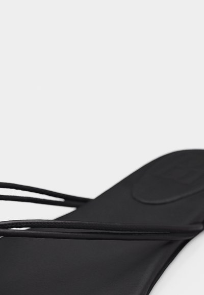 Κοντινό πλάνο μαύρου σανδαλιού που δείχνει λεπτά λουράκια και λεία επιφάνεια πέλματος με ουδέτερο φόντο.