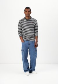 Svart-hvitt stripete langermet skjorte med en liten logo, kombinert med vide blå denim cargo-bukser med sidelommer og hvite joggesko.