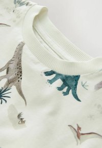 Baumwollhemd mit Rundhalsausschnitt, mit hellem cremefarbenem Hintergrund und bunten Dinosaurierillustrationen in Grün, Braun und Blau.