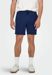 Pantalones cortos azul marino hechos de tejido texturizado con cinturilla elástica y cordón ajustable. Llevado con calcetines blancos y zapatillas.