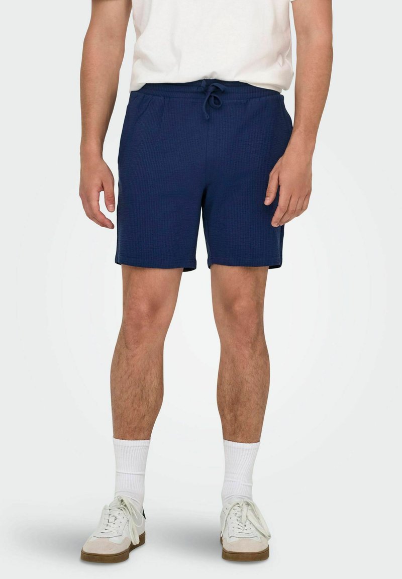 Pantalones cortos azul marino hechos de tejido texturizado con cinturilla elástica y cordón ajustable. Llevado con calcetines blancos y zapatillas.