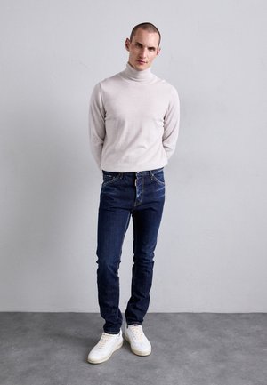 Mann, der gegen eine hellgraue Wand steht, gekleidet in einem beigen Rollkragenpullover, dunkelblauen Jeans und weißen Turnschuhen, mit den Händen hinter dem Rücken.