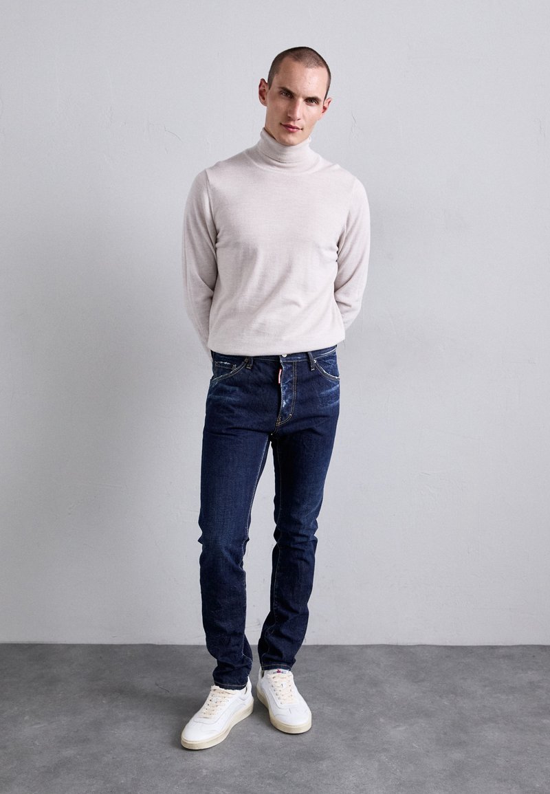 Mann, der gegen eine hellgraue Wand steht, gekleidet in einem beigen Rollkragenpullover, dunkelblauen Jeans und weißen Turnschuhen, mit den Händen hinter dem Rücken.