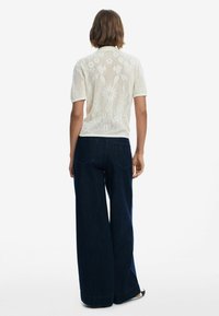 Polo in maglia crema a maniche corte con motivo floreale a struttura aperta sulla schiena, abbinato a jeans palazzo blu scuro. Calzature semplici visibili.